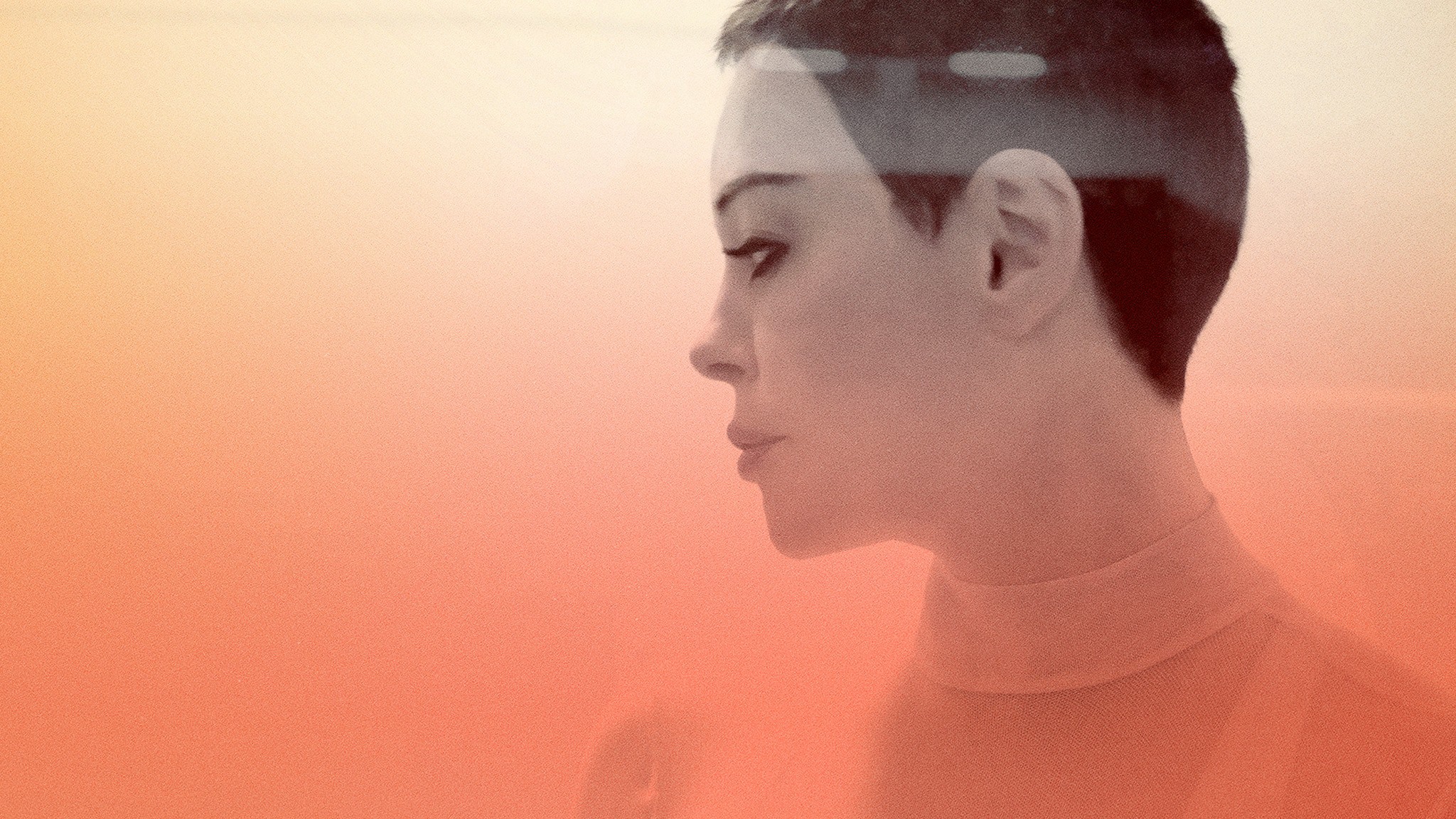 A medium closeup side-profile shot of Rose McGowan. Her face is desaturated and a peach-colored filter spreads from the bottom of the image.